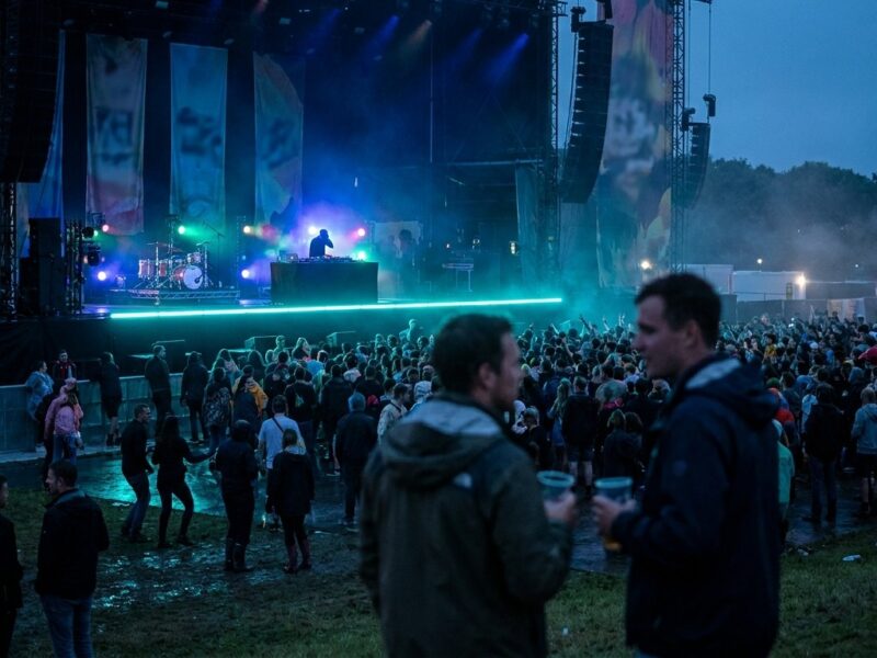 Festivalbühne bei Nacht mit Lichtshow, Crowd auf nasser Wiese und zwei Besuchern im Vordergrund