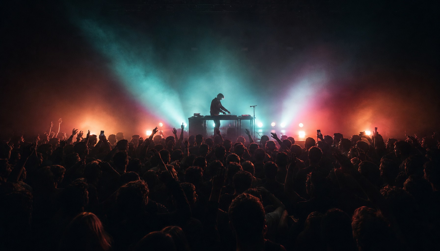 DJ steht auf einer Konzertbuehne vor dichtem Publikum, umgeben von Nebel und farbigen Scheinwerfern.