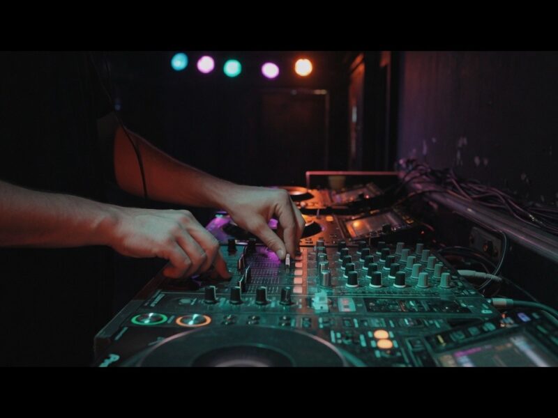 Hände bedienen ein DJ-Mischpult mit Reglern, Kabeln und bunten Lichtern in dunkler Clubumgebung