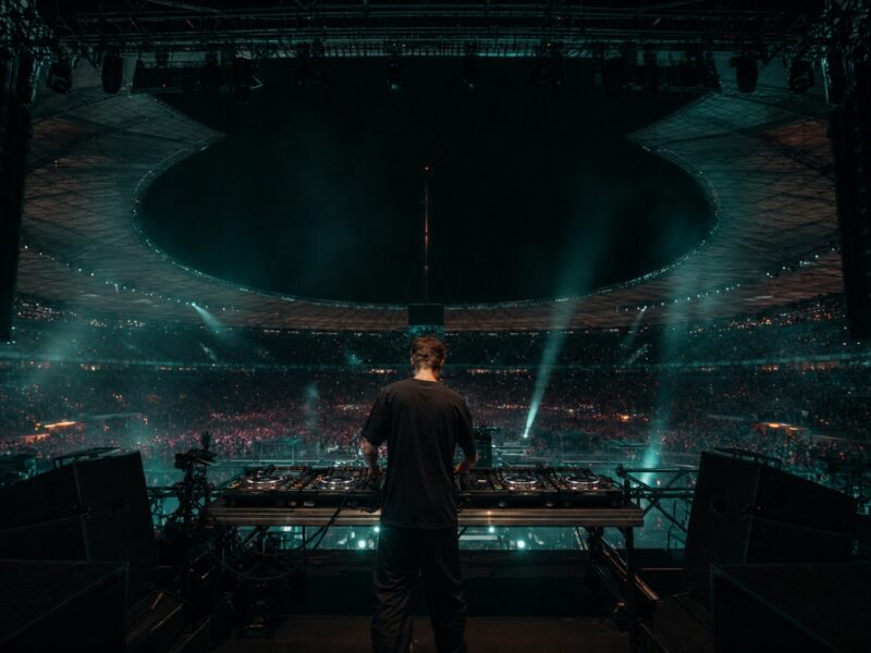 DJ steht mit Mischpult auf einer Stadionbühne vor dichtem Publikum und beleuchteten Rängen