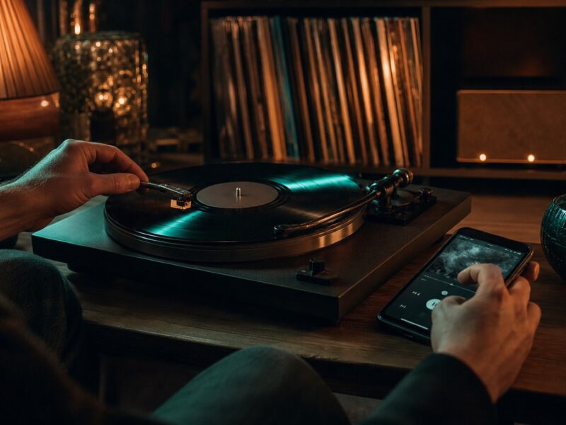 Hand legt Tonarm auf eine Schallplatte, daneben steuert eine zweite Hand Musik am Smartphone.